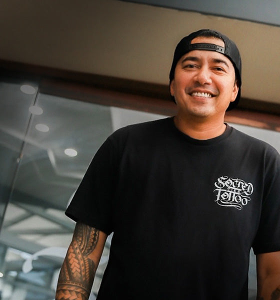 Smiling person in a black cap and T-shirt with a tattoo on their arm.
