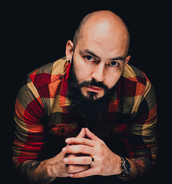 Bald man with a beard wearing a plaid shirt, leaning forward with clasped hands.