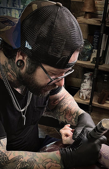 Tattoo artist wearing a cap and gloves, focused on tattooing.