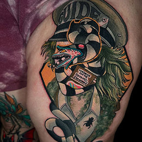 Tattoo of a surreal figure with a snake tongue and text elements on upper arm.